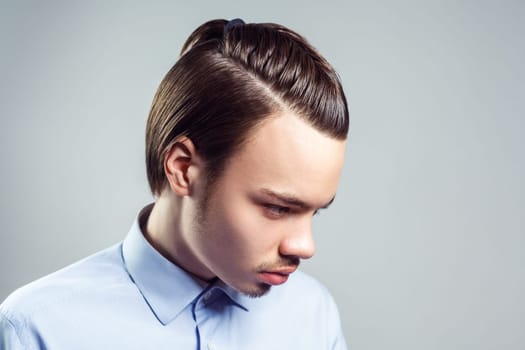 Side view portrait of young adult brunette man with mustache and beard with top knot hairstyle, looking away, wearing blue shirt. Indoor studio shot isolated on gray background.