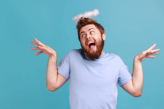 Portrait of handsome funny bearded angelic man with holy nimbus on head standing with raised hands and looking away with open mouth. Indoor studio shot isolated on blue background.