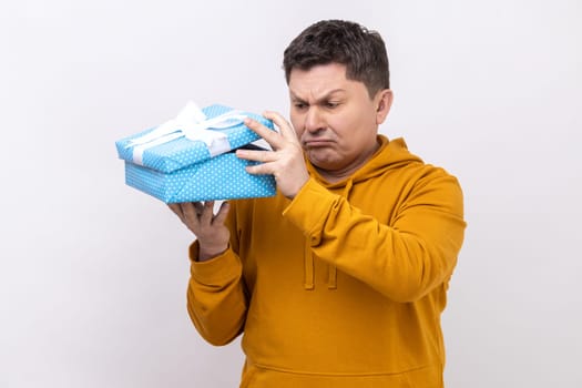 Portrait of unsatisfied displeased man holding in hands unpacked gift box, frustrated with content, bad present, wearing urban style hoodie. Indoor studio shot isolated on white background.