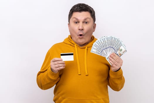 Shocked middle aged man holding and showing dollar cash and credit card, surprised with cashback, currency exchange, wearing urban style hoodie. Indoor studio shot isolated on white background.