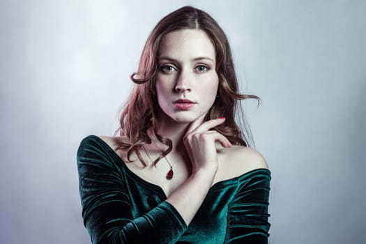 Beauty portrait of self-confident attractive woman with freckles wearing green dress holding her chin and looking ar camera with serious expression. Indoor studio shot isolated on gray background.