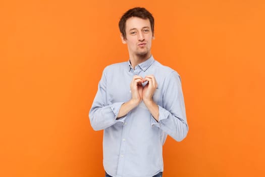 Portrait of funny romantic handsome man standing and showing heart shape gesture, falling in love, wearing light blue shirt. Indoor studio shot isolated on orange background.