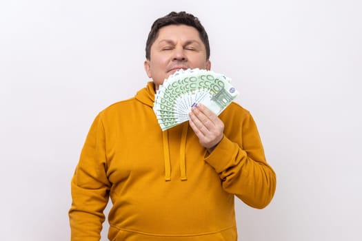 Greedy mercantile man smelling fan of cash, enjoying aroma of hundred euro bills in his hand, satisfied with big profit, wearing urban style hoodie. Indoor studio shot isolated on white background.