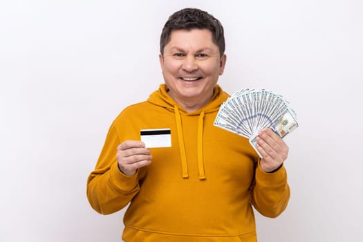 Happy middle aged man holding and showing dollar cash and credit card, satisfied with cashback, currency exchange, wearing urban style hoodie. Indoor studio shot isolated on white background.