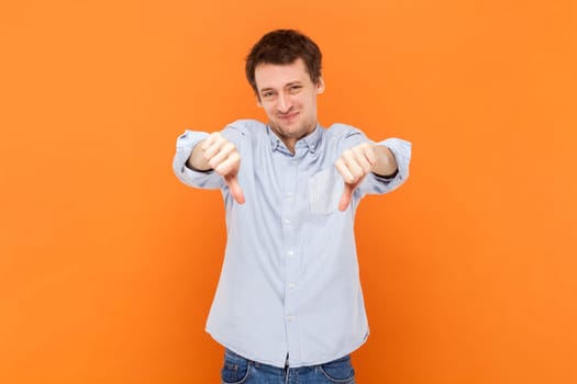 Portrait of sad upset handsome man demonstrates disapproval gesture, dislike service, showing thumbs down, wearing light blue shirt. Indoor studio shot isolated on orange background.