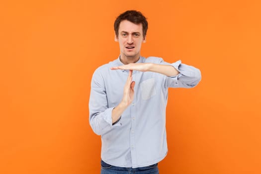 Portrait of serious self confident man making timeout gesture, demonstrating limit, asking to stop, wearing light blue shirt. Indoor studio shot isolated on orange background.