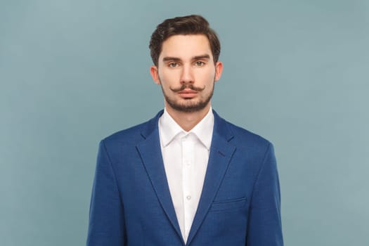 Portrait of serious strict bossy man with mustache standing looking at camera, being in bad mood, wearing official style suit. Indoor studio shot isolated on light blue background.