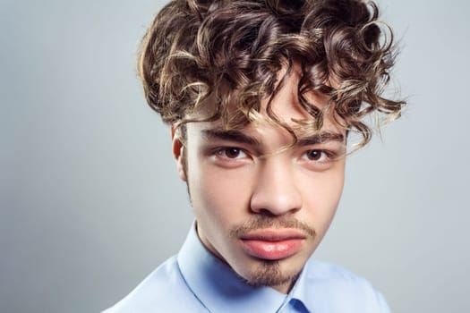 Portrait of handsome man with mustache and beard with curly hairstyle, wearing blue shirt, looking at camera with serious expression. Indoor studio shot isolated on gray background.