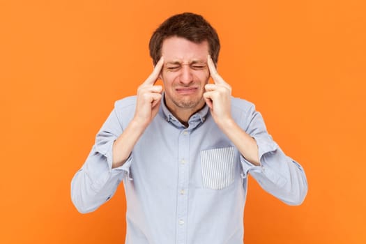 Portrait of unhealthy sad sick man standing with fingers near temples, having migraine, frowning face, wearing light blue shirt. Indoor studio shot isolated on orange background.