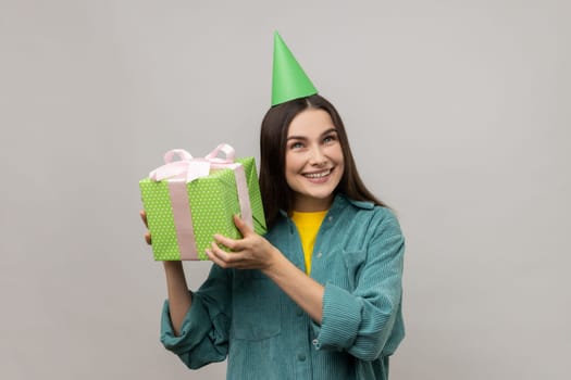 Happy smiling woman in party cap holding and shaking green gift box trying to guess what is inside, birthday surprise, wearing casual style jacket. Indoor studio shot isolated on gray background.