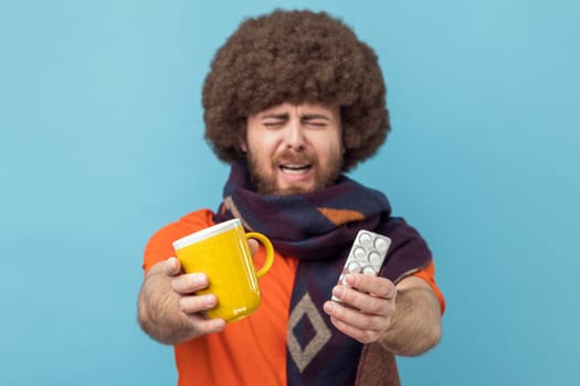 Portrait of man with Afro hairstyle wearing orange T-shirt and wrapped in scarf, treating with pills and warm tea, being upset, feeling unwell. Indoor studio shot isolated on blue background.
