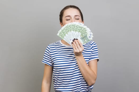 Portrait of avaricious woman wearing striped T-shirt smelling euro banknotes with expression of pleasure, enjoying financial success, wealthy life. Indoor studio shot isolated on gray background.