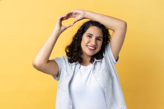 Portrait of woman with dark wavy hair standing with raised hands and making heart or love gesture and looking at camera with toothy smile. Indoor studio shot isolated on yellow background.