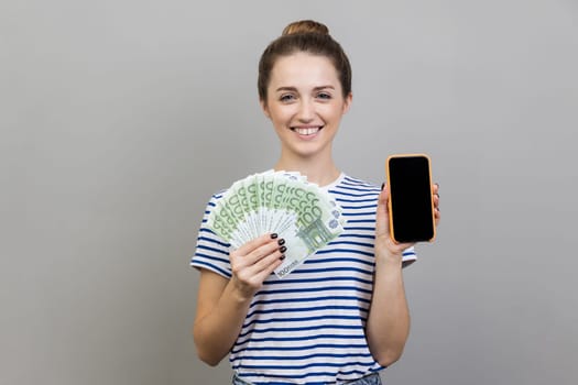 Portrait of woman wearing striped T-shirt holding fan of euro banknotes and smart phone with black blank screen, making money online. Indoor studio shot isolated on gray background.