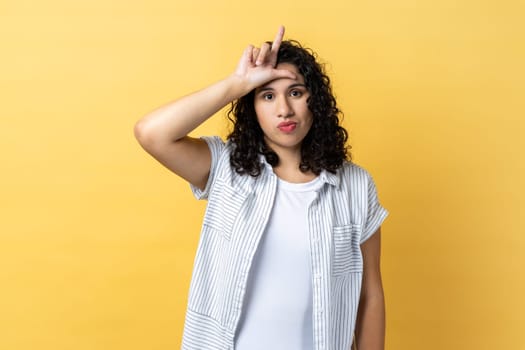 Portrait of depressed woman with dark wavy hair holding hand near forehead showing loser gesture with fingers, having problems, looks unhappy. Indoor studio shot isolated on yellow background.