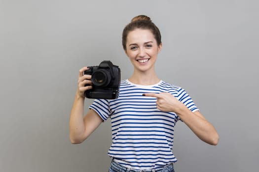 Portrait of woman photographer wearing striped T-shirt pointing at professional digital dslr camera and looking with toothy smile, enjoying her job. Indoor studio shot isolated on gray background.