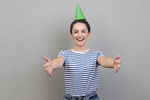 Come into my arms. Portrait of pleased smiling woman wearing striped T-shirt and party cone, inviting her friends to birthday party. Indoor studio shot isolated on gray background.