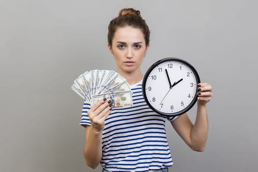 Portrait of strict serious woman with bun hairstyle wearing striped T-shirt holding wall clock and fun of dollar banknotes, looking at camera. Indoor studio shot isolated on gray background.