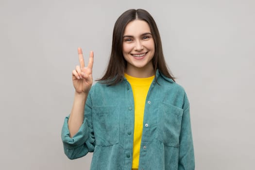 Portrait of happy attractive excited woman gesturing victory sign at camera, success or achievement, expressing happiness, wearing casual style jacket. Indoor studio shot isolated on gray background.