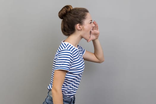 Side view of woman wearing striped T-shirt keeps arms around wide open mouth, screaming loudly, calling attention to important news, announcing event. Indoor studio shot isolated on gray background.