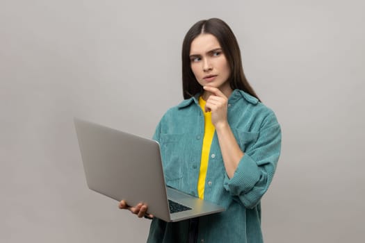 Portrait of pensive thoughtful smart dark haired woman holding laptop and thinking about school project, wearing casual style jacket. Indoor studio shot isolated on gray background.