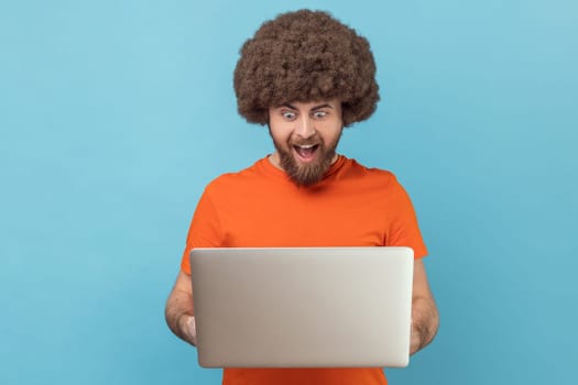 Portrait of excited amazed man with Afro hairstyle wearing orange T-shirt holding notebook and looking at display with excitement, playing computer games Indoor studio shot isolated on blue background