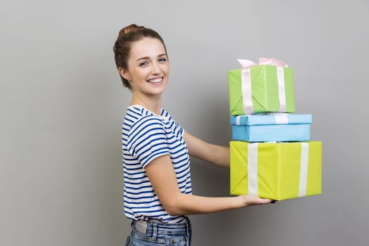 Portrait of cheerful woman wearing striped T-shirt giving stack of presents, looking at camera, giving gifts to her friend, having festive mood. Indoor studio shot isolated on gray background.