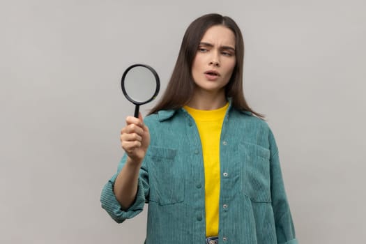 Curious woman with dark hair looking away through magnifying glass, spying, finding out something, inspecting, wearing casual style jacket. Indoor studio shot isolated on gray background.