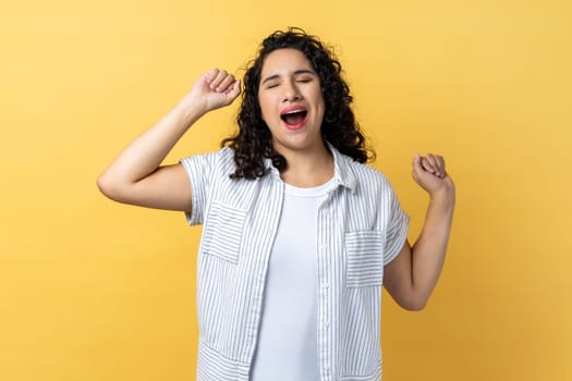 Portrait of tired sleepy beautiful woman with dark wavy hair yawning and raising hands up, feeling fatigued, standing with close eyes. Indoor studio shot isolated on yellow background.