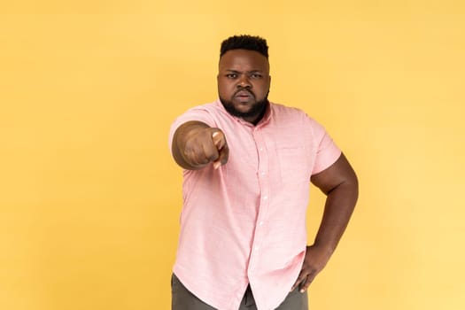 Portrait of bearded young adult man wearing pink shirt noticing and pointing finger to camera, accusing with serious bossy face, making choice. Indoor studio shot isolated on yellow background.