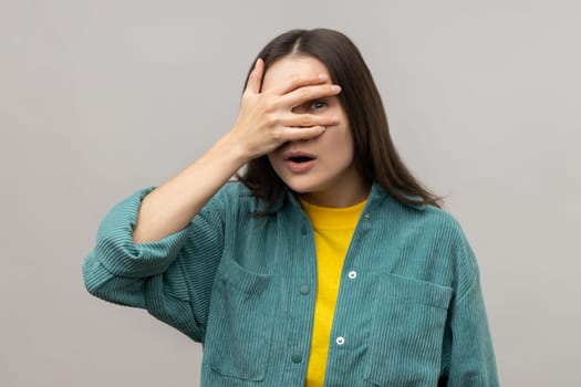 Portrait of curious dark haired woman spying, hiding and peeping through fingers, looking for secrets, rumors, wearing casual style jacket. Indoor studio shot isolated on gray background.