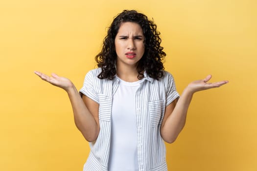 What do you want. Portrait of woman with dark wavy hair standing with raised hands and surprised indignant expression, asking what reason. Indoor studio shot isolated on yellow background.