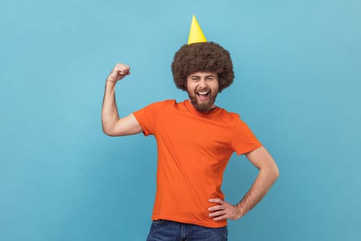 Portrait of strong powerful man with Afro hairstyle wearing orange T-shirt and party cone standing with raised arm, showing his biceps. Indoor studio shot isolated on blue background.