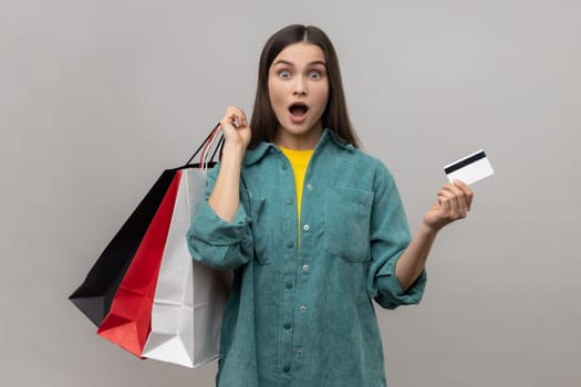 Surprised woman holding and showing limitless credit card and shopping bags, shocked with prices, final sale, wearing casual style jacket. Indoor studio shot isolated on gray background.