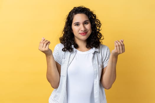 Give me money, many dollars. Woman with dark wavy hair showing money gesture, hinting at salary increase, having business idea to earn income. Indoor studio shot isolated on yellow background.