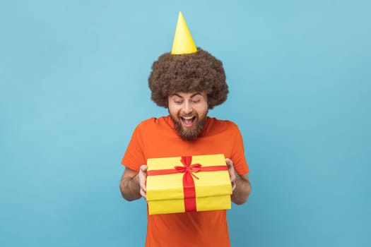 Portrait of happy man with Afro hairstyle wearing orange T-shirt holding gift and looking at camera with toothy smile, gifts and bonuses, celebration. Indoor studio shot isolated on blue background.