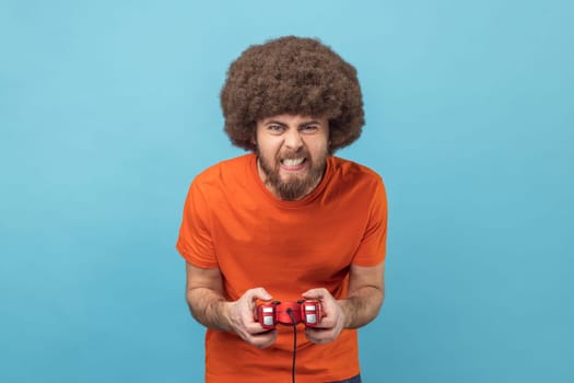 Portrait of crazy funny man with Afro hairstyle grimacing holding joystick, playing video games online, virtual competition. Indoor studio shot isolated on blue background.