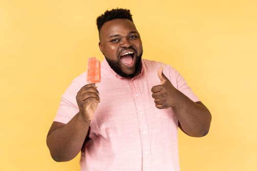 Portrait of excited happy satisfied bearded man wearing pink holding ice cream, showing thumb up, likes tasty cold dessert. shirt Indoor studio shot isolated on yellow background.