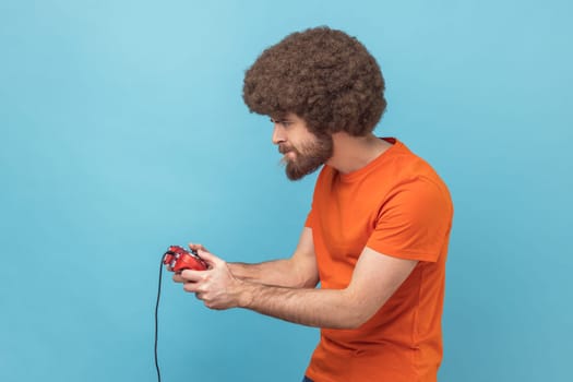 Side view of concentrated man with Afro hairstyle wearing orange T-shirt holding in hands red gamepad joystick, grimacing playing video games. Indoor studio shot isolated on blue background.