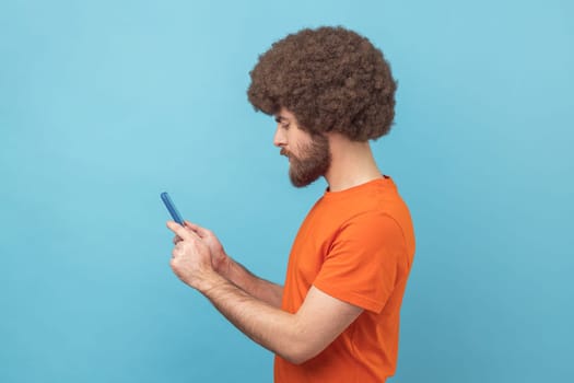 Side view of concentrated man with Afro hairstyle wearing orange T-shirt writing post on social network using mobile phone, chatting looking serious. Indoor studio shot isolated on blue background.