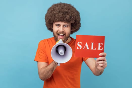 Portrait of smiling man with Afro hairstyle wearing orange T-shirt holding card with sale inscription and megaphone, telling about discounts. Indoor studio shot isolated on blue background.