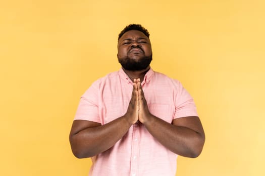 Portrait of unhappy man wearing pink shirt looking up with beseeching imploring expression, praying and asking heartily, feeling desperate urging help. Indoor studio shot isolated on yellow background