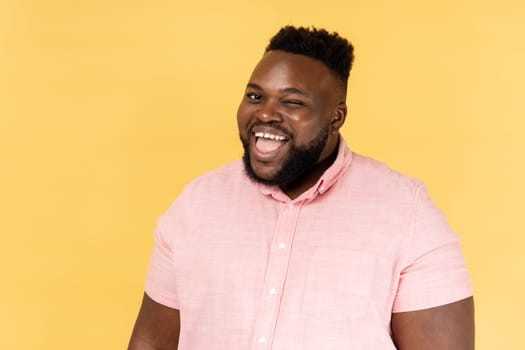 Portrait of childish happy young adult man wearing pink shirt winking and optimistically smiling, good mood, cunning bearded guy flirting. Indoor studio shot isolated on yellow background.