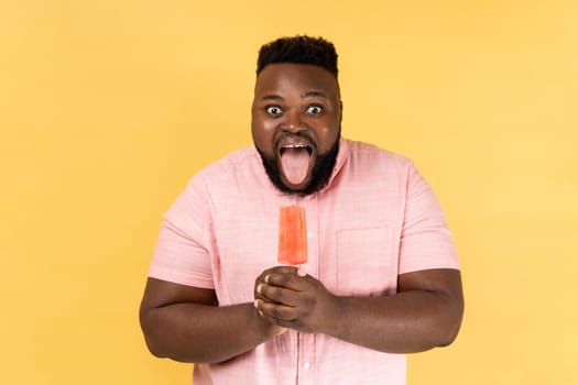 Portrait of funny positive bearded young adult man wearing pink shirt holding pink cold ice cream and showing tongue out, feels hungry. Indoor studio shot isolated on yellow background.