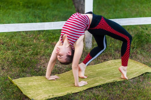 Full length of young adult attractive yogi woman practicing yoga, stretching in wheel exercise, urdhva dhanurasana pose, working out on mat outdoor in green park, wearing sportswear.