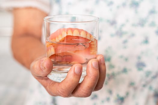 Asian senior woman patient holding and washing denture in water cleanser glass for good chewing.