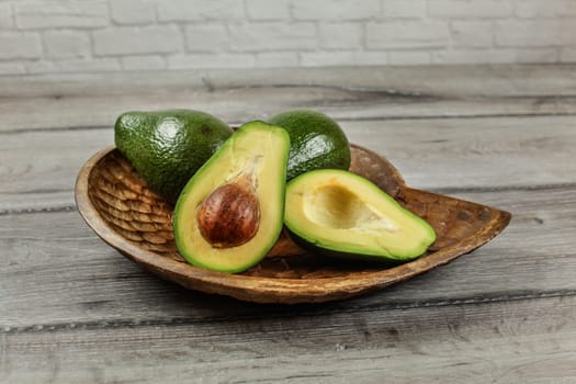 Avocado cut in half, seed and pulp visible, two more whole pears on carved wooden bowl.