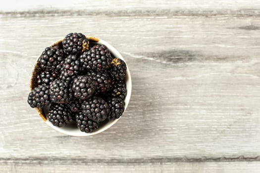Tabletop view, small ceramic cup with blackberries on gray wood desk. Space for text on the right.