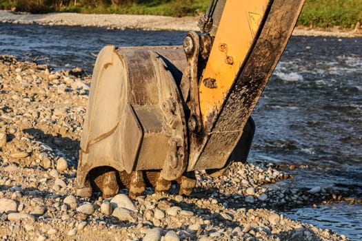 Digger (excavator machine) bucket digging stones ground by the river.