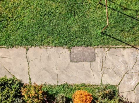 Old cracked pavement with green lawn and small bushes around. View from above, balcony on high floor flat.